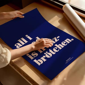 May include: A rectangular dark blue poster featuring white text that reads all i need is br&ouml;tchen. The design includes the smaller text design by frost & freckles in the bottom right corner. The poster is displayed on a flat surface.