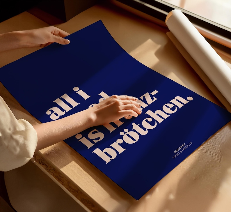 May include: A rectangular dark blue poster featuring white text that reads all i need is br&ouml;tchen. The design includes the smaller text design by frost & freckles in the bottom right corner. The poster is displayed on a flat surface.