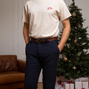 May include: A cream-coloured short-sleeve t-shirt featuring the red text Merry Christmas embroidered on the upper left chest, paired with dark navy blue trousers and a brown leather belt with a rectangular metal buckle at the waist.