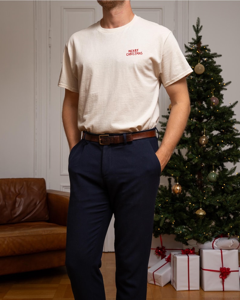 May include: A cream-coloured short-sleeve t-shirt featuring the red text Merry Christmas embroidered on the upper left chest, paired with dark navy blue trousers and a brown leather belt with a rectangular metal buckle at the waist.