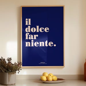May include: A rectangular wall art print in a light wood frame featuring a deep blue background with the white text "il dolce far niente." and the smaller text "DESIGN BY FROST & FRECKLES" at the bottom, displayed in a clean, modern interior space.