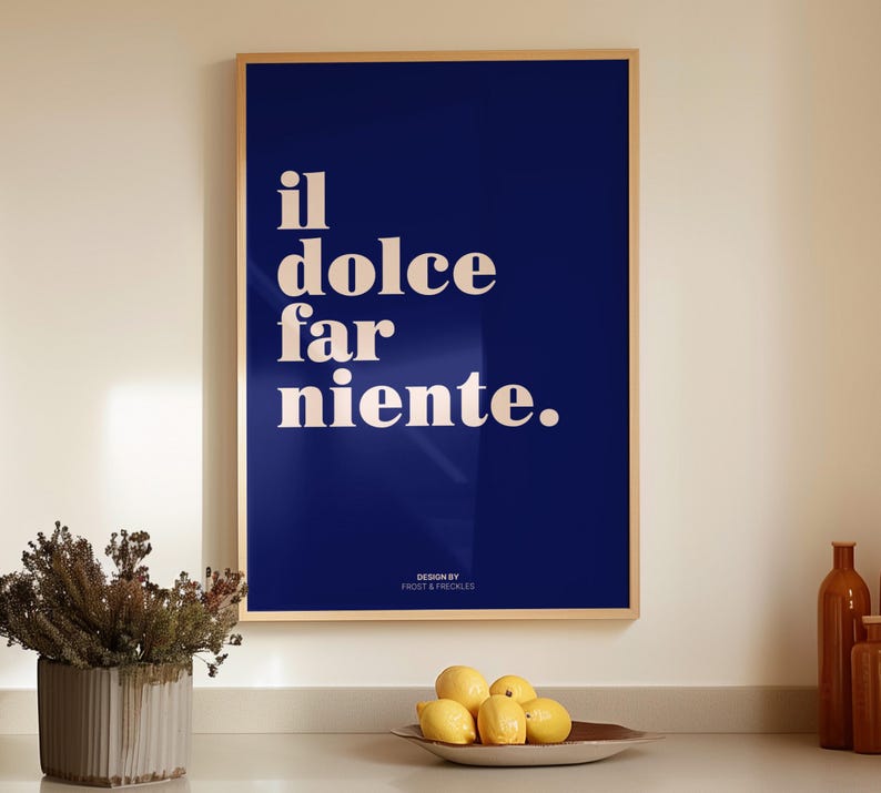 May include: A rectangular wall art print in a light wood frame featuring a deep blue background with the white text "il dolce far niente." and the smaller text "DESIGN BY FROST & FRECKLES" at the bottom, displayed in a clean, modern interior space.