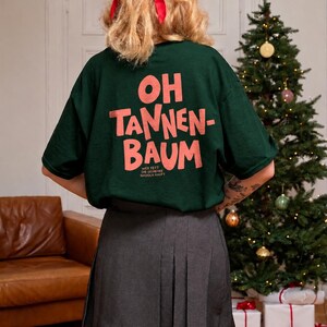 May include: A dark green short-sleeved t-shirt featuring the pink text "OH TANNEN-BAUM" and smaller text "WER FEET DIE GESCHENKE NADOLN KAUFT" on the back, paired with a grey pleated skirt and a red hair ribbon.