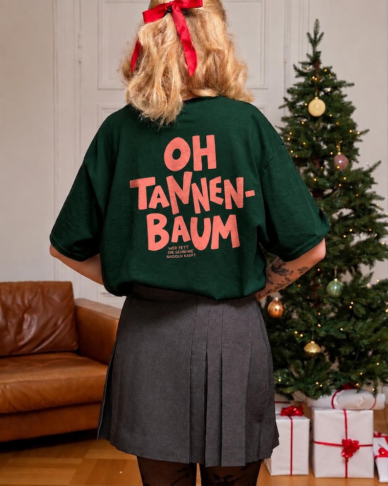 May include: A dark green short-sleeved t-shirt featuring the pink text "OH TANNEN-BAUM" and smaller text "WER FEET DIE GESCHENKE NADOLN KAUFT" on the back, paired with a grey pleated skirt and a red hair ribbon.