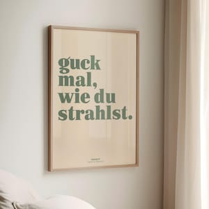 May include: A framed poster with the text "guck mal, wie du strahlst" in green letters on a beige background. The poster is in a light brown wooden frame and is hanging on a white wall.