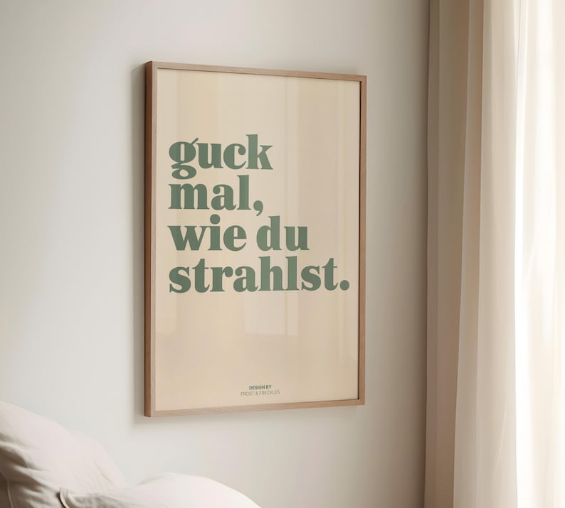 May include: A framed poster with the text "guck mal, wie du strahlst" in green letters on a beige background. The poster is in a light brown wooden frame and is hanging on a white wall.