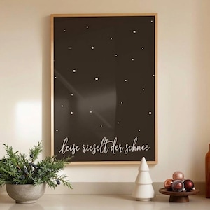 May include: A rectangular wall art print in a light wood frame featuring a dark brown background with scattered white dots resembling falling snow and the white cursive text leise rieselt der schnee at the bottom of the print.