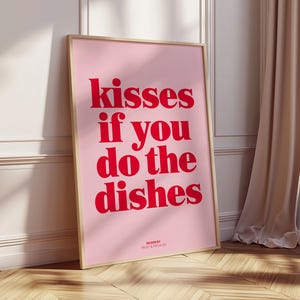 May include: A framed pink poster with the text "kisses if you do the dishes" in red. The poster is leaning against a white wall and a wooden floor. The words "DESIGN BY" and a name are at the bottom.