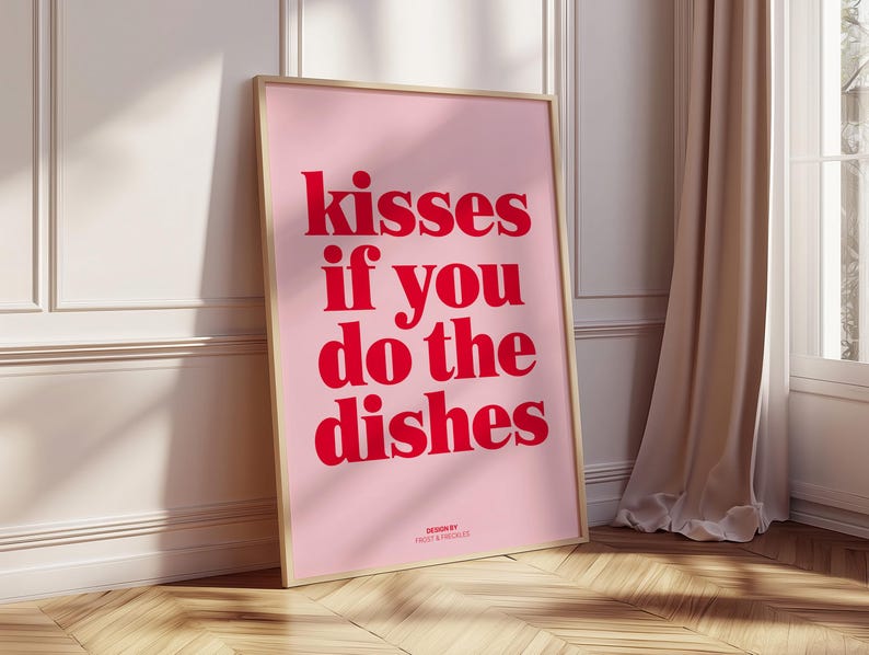 May include: A framed pink poster with the text "kisses if you do the dishes" in red. The poster is leaning against a white wall and a wooden floor. The words "DESIGN BY" and a name are at the bottom.