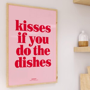 May include: A framed pink poster with the text "kisses if you do the dishes" in red. The poster has a light wood frame and is displayed on a white wall. The words "DESIGN BY FROST & FRECKLES" are at the bottom.