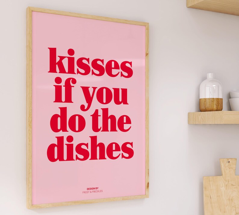 May include: A framed pink poster with the text "kisses if you do the dishes" in red. The poster has a light wood frame and is displayed on a white wall. The words "DESIGN BY FROST & FRECKLES" are at the bottom.