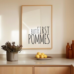 Może przedstawiać: Wydruk w ramie z tekstem "But first, pommes" czarnymi literami na białym tle. Wydruk wisi na białej ścianie nad blatem z miską cytryn.