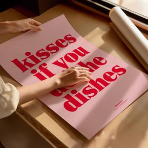 May include: A pink poster with the text "kisses if you do the dishes" in bold red letters. The poster is on a wooden surface, with a rolled-up white poster in the background. The design is by Frost & Freckles.