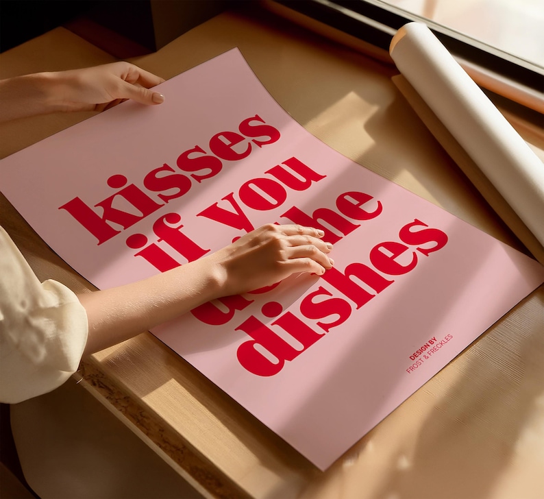May include: A pink poster with the text "kisses if you do the dishes" in bold red letters. The poster is on a wooden surface, with a rolled-up white poster in the background. The design is by Frost & Freckles.