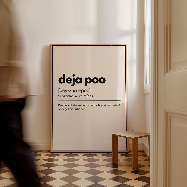 May include: A framed art print with the words "deja poo" in a bold, black font. The print includes a phonetic pronunciation and a German definition. A small wooden side table sits nearby on a checkered floor.