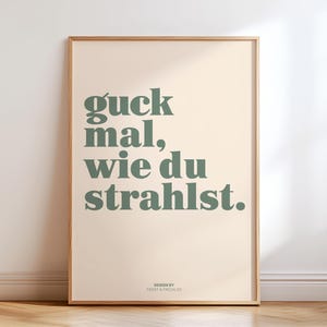May include: A framed art print with the German phrase "guck mal, wie du strahlst" in green text on a cream background. The phrase translates to "look how you shine." The design is by Frost & Freckles.