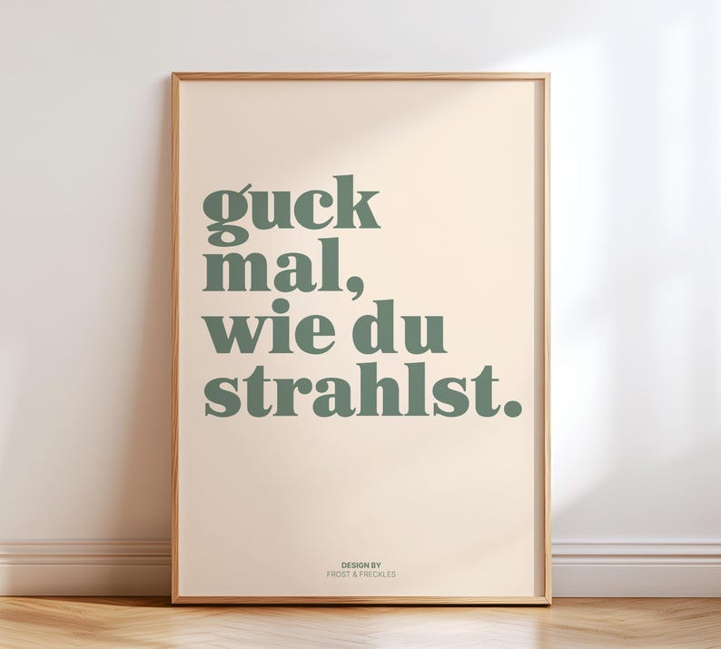 May include: A framed art print with the German phrase "guck mal, wie du strahlst" in green text on a cream background. The phrase translates to "look how you shine." The design is by Frost & Freckles.
