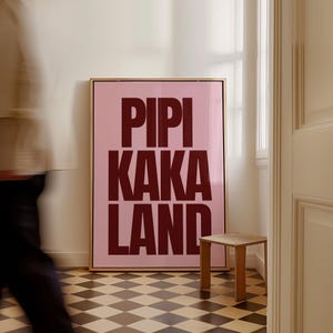 Peut inclure: Une affiche rose encadr&eacute;e avec les mots "PIPI KAKA LAND" en lettres majuscules rouge fonc&eacute;. L'&oelig;uvre est expos&eacute;e dans une pi&egrave;ce avec un sol &agrave; carreaux et une petite table d'appoint en bois.