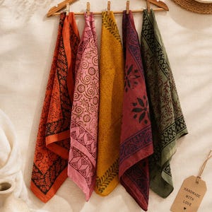 May include: Five square fabric napkins hanging from a wooden hanger with small clips. The napkins feature distinct patterns in orange, pink, mustard yellow, maroon, and olive green. A tan tag attached to the side reads HANDMADE WITH LOVE.