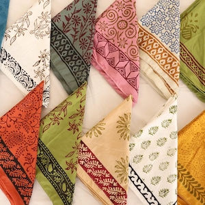 May include: A collection of twelve triangular folded fabric napkins arranged in two rows. Each napkin features a unique block-printed pattern in various colors including blue, white, grey, pink, green, orange, yellow, and maroon with floral motifs.