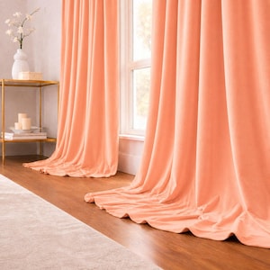 Op de afbeelding: Perzikroze fluwelen gordijnen draperen voor een raam. De gordijnen, die tot de vloer reiken, hebben zachte plooien en een subtiele glans. Een gouden bijzettafel met een witte vaas en kaarsen is ook zichtbaar.