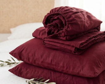 May include: A stack of deep burgundy linen bedding consisting of two rectangular pillows and a folded sheet. The fabric has a visible natural texture and is accented by small green olive branches placed on the surface.