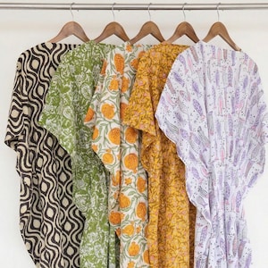 May include: Five colorful, patterned caftans hanging on a wooden rod. The caftans feature various designs, including geometric shapes, floral patterns, and fish motifs. Colors include brown, green, yellow, and purple. The caftans are made of lightweight fabric.