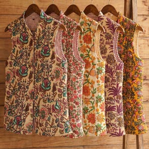 Puede incluir: Seis chalecos acolchados sin mangas con estampados florales en crema, rojo, naranja y amarillo. Cuelgan de perchas de madera sobre un fondo de madera.