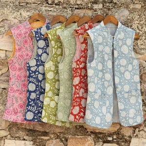 Puede incluir: Una colección de chalecos sin mangas en varios colores, cada uno adornado con un estampado floral. Los chalecos se exhiben en perchas de madera contra una pared de piedra texturizada. Los colores incluyen rosa, azul marino, verde, rojo y azul claro.