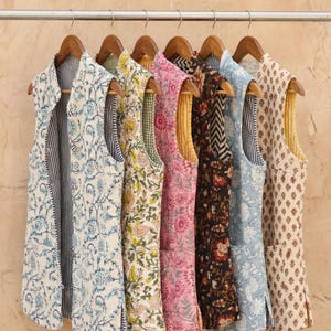 May include: Seven sleeveless vests with diverse floral and geometric patterns hang on a metal rail with wooden hangers. The vests display colours including blue, pink, yellow, and black, with contrasting interior linings.