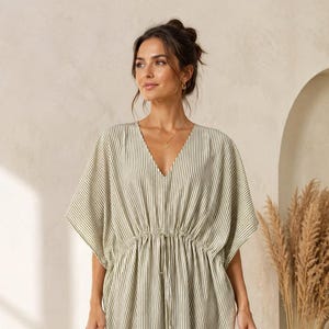 May include: A floor-length kaftan dress featuring a vertical olive green and white striped pattern. The garment has a V-neckline, wide kimono-style sleeves, and a drawstring waist with thin ties that hang down the center front of the dress.