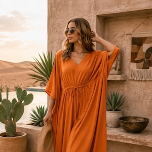 May include: A solid burnt orange midi-length kaftan dress featuring a V-neckline, loose elbow-length sleeves, and a drawstring waist tie. The garment is made of a smooth, lightweight fabric that drapes loosely down to the mid-calf area.