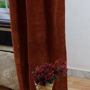 May include: A rust-colored velvet curtain hangs in front of a white wall. A wicker vase with a bouquet of dark red flowers sits on the wooden floor in front of the curtain.