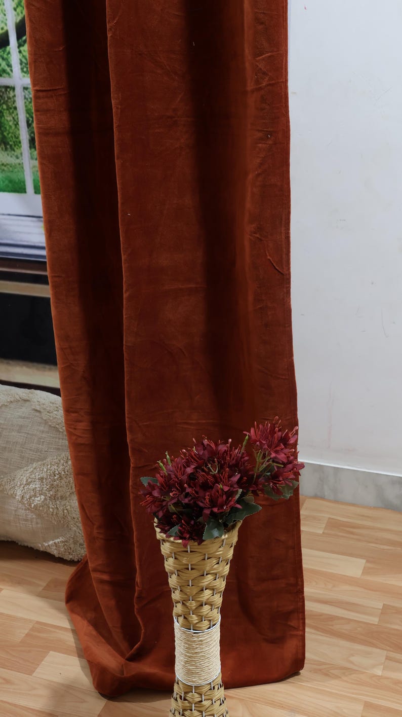 May include: A rust-colored velvet curtain hangs in front of a white wall. A wicker vase with a bouquet of dark red flowers sits on the wooden floor in front of the curtain.