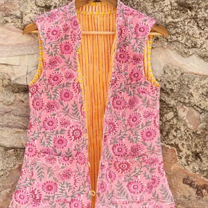 May include: A sleeveless pink floral print vest with a yellow and orange striped lining. The vest is hanging on a wooden hanger against a textured stone wall. The floral pattern includes pink flowers and gray leaves.