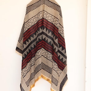 May include: A patterned scarf with a geometric design in black, red, and beige. The scarf has a gold border and is hanging against a white background. The design includes stripes, zigzags, and small geometric shapes.