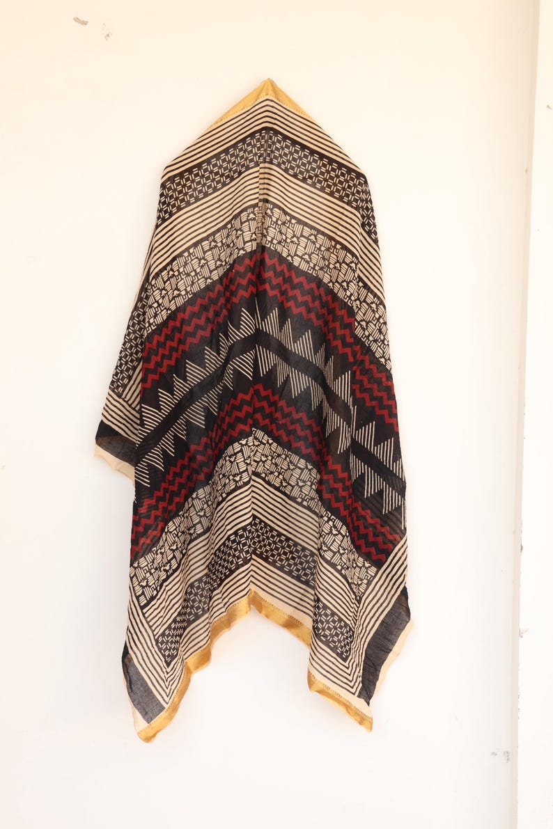 May include: A patterned scarf with a geometric design in black, red, and beige. The scarf has a gold border and is hanging against a white background. The design includes stripes, zigzags, and small geometric shapes.
