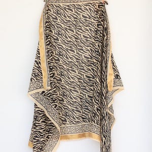 May include: A black and beige patterned shawl hanging on a wooden hanger. The shawl features a zebra stripe design with a beige border and geometric patterns. The fabric appears lightweight and flowing.