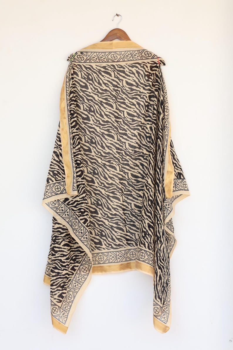 May include: A black and beige patterned shawl hanging on a wooden hanger. The shawl features a zebra stripe design with a beige border and geometric patterns. The fabric appears lightweight and flowing.