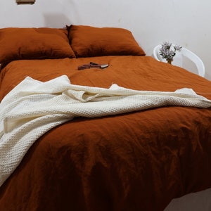 Könnte beinhalten: Ein rostfarbener Leinen-Bettbezug mit einer weißen Waffel-Decke darauf. Das Bett ist mit zwei rostfarbenen Leinen-Kissenbezügen bezogen.