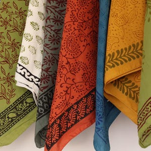 May include: A collection of hanging fabric squares featuring diverse block-printed patterns in shades of maroon, olive green, cream, orange, blue, yellow, and pink. Each square displays intricate floral, paisley, and geometric motifs in contrasting ink.