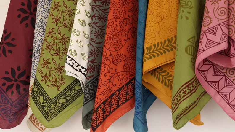 May include: A collection of hanging fabric squares featuring diverse block-printed patterns in shades of maroon, olive green, cream, orange, blue, yellow, and pink. Each square displays intricate floral, paisley, and geometric motifs in contrasting ink.