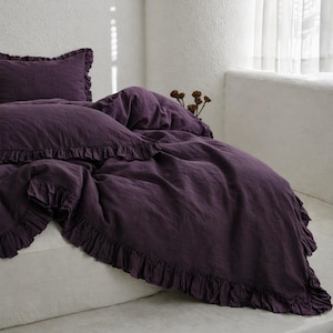 May include: A deep purple bedding set featuring a ruffled duvet cover and pillowcase. The set is made of a soft, textured fabric and is displayed on a white bed. The ruffled edges add a decorative touch to the bedroom decor.