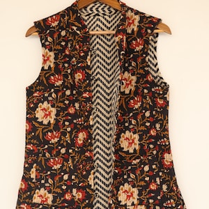 May include: A sleeveless vest with a floral pattern in red, cream, and gold on a black background. The interior lining features a black and cream chevron design. The vest is hanging on a wooden hanger.