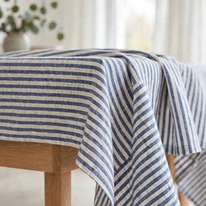 May include: A rectangular tablecloth featuring a pattern of vertical navy blue and white stripes. The fabric has a visible woven texture and drapes over the edges of a wooden table, covering the top surface and hanging down on the sides.