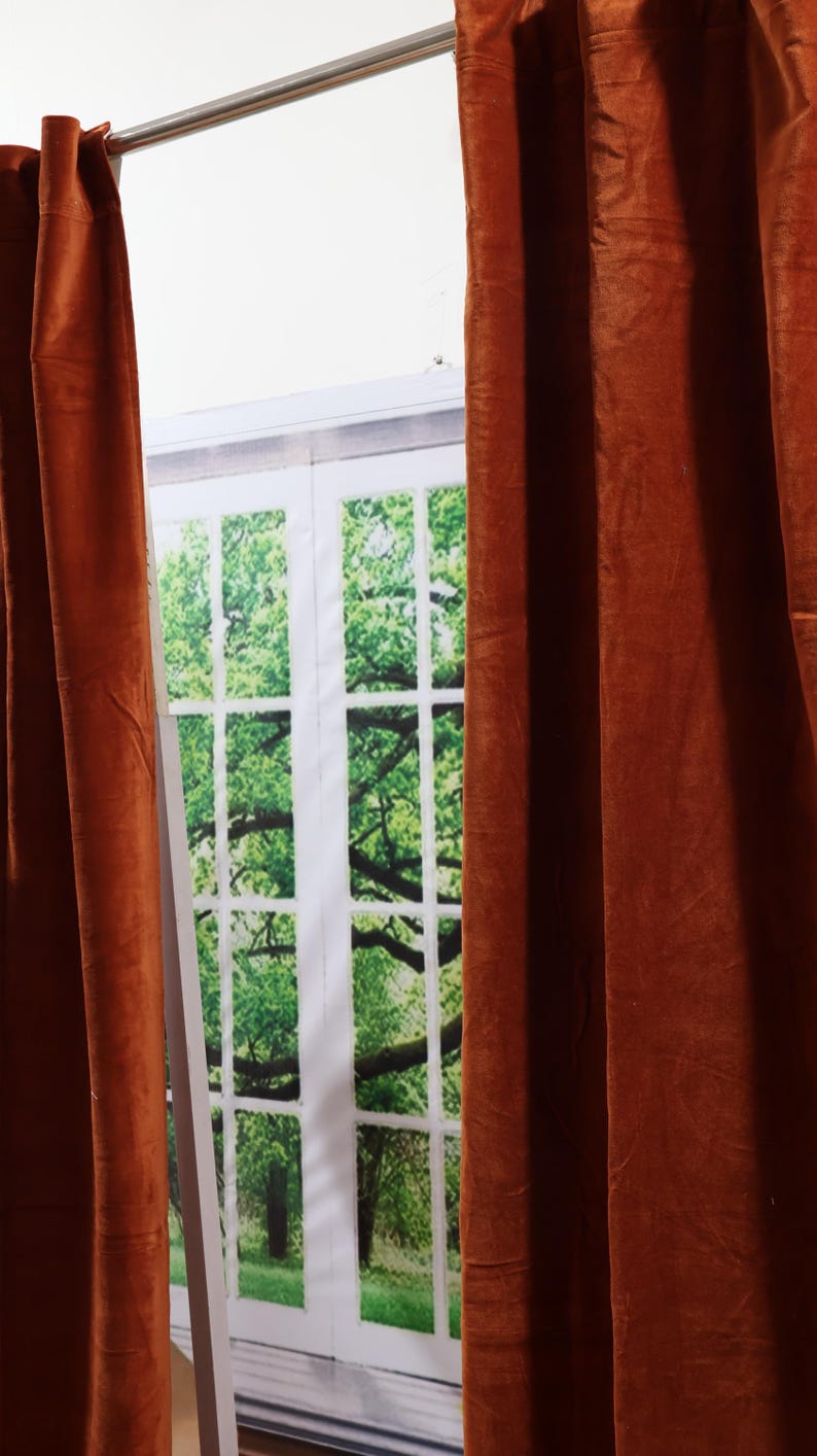 May include: Two rust-colored velvet curtains hang in front of a window with a printed image of a green tree and a white frame.
