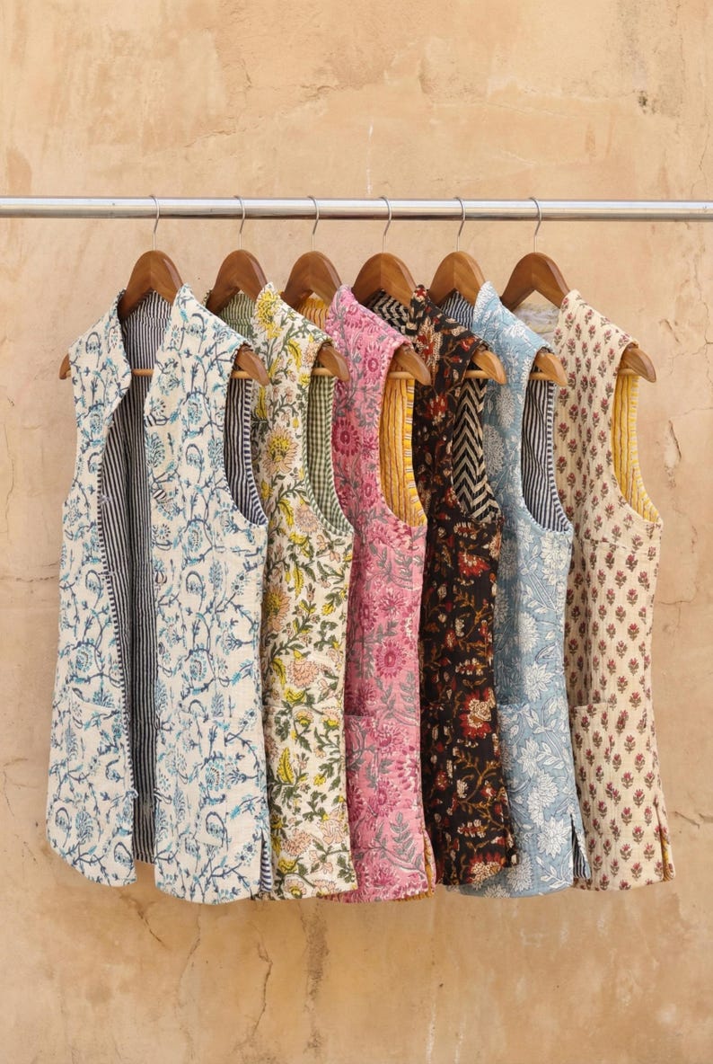 May include: A collection of seven sleeveless vests with various floral and geometric patterns. The vests hang on a metal rod with wooden hangers. The vests feature colors like blue, pink, yellow, and black, with contrasting interior linings.