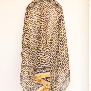 May include: A beige scarf with a repeating pattern of black fish. The scarf has a decorative border with floral and geometric designs. The edges are trimmed with a gold-colored fabric.
