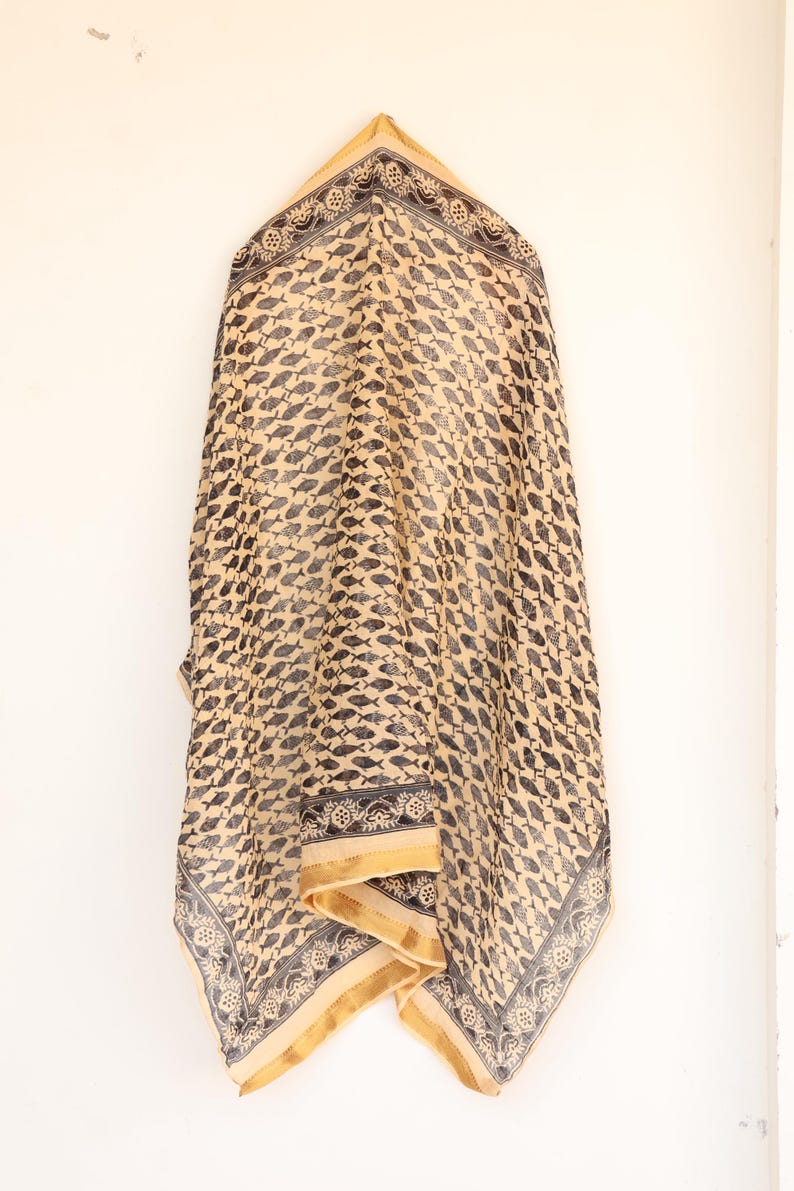 May include: A beige scarf with a repeating pattern of black fish. The scarf has a decorative border with floral and geometric designs. The edges are trimmed with a gold-colored fabric.