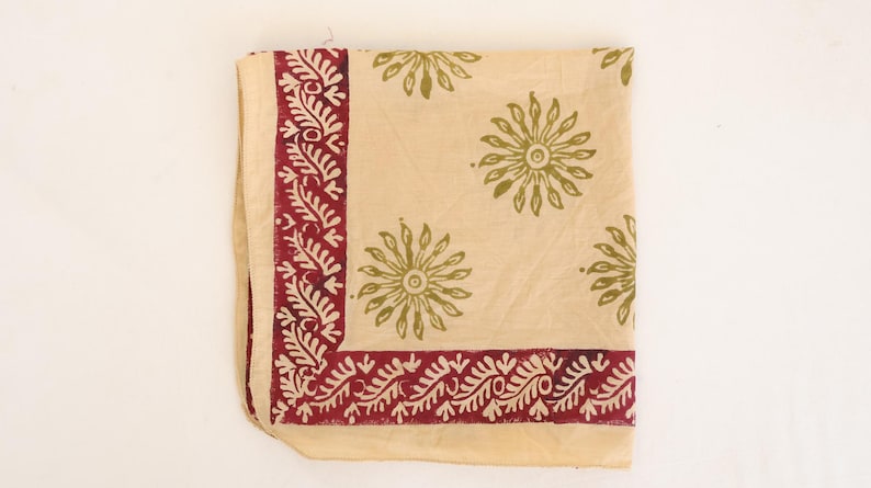 May include: A square textile piece in a light beige color featuring a dark red border with a repeating leaf pattern. The center of the fabric is decorated with a scattered print of olive green sunburst or floral motifs across the surface.